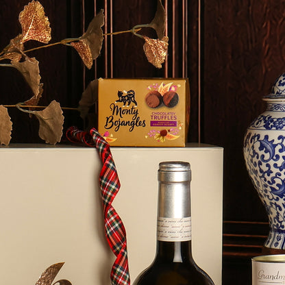 A box of Monty Bojangles chocolate truffles displayed on a white gift box next to a bottle of wine, with a red plaid ribbon, decorative dried leaves, and a blue-and-white ceramic vase in the background.