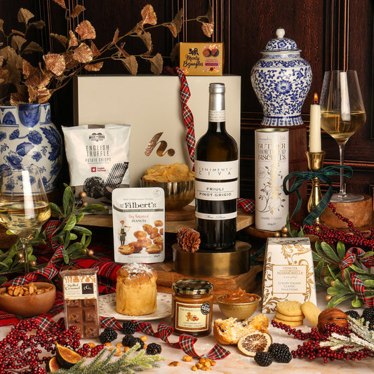The Let It Snow Christmas gift hamper arranged on a tabletop with a bottle of white wine, a pack of English Truffle crisps, a box of chocolate truffles, a small panettone, biscuits, nuts, a candle, and decorative festive elements against a dark wood background.