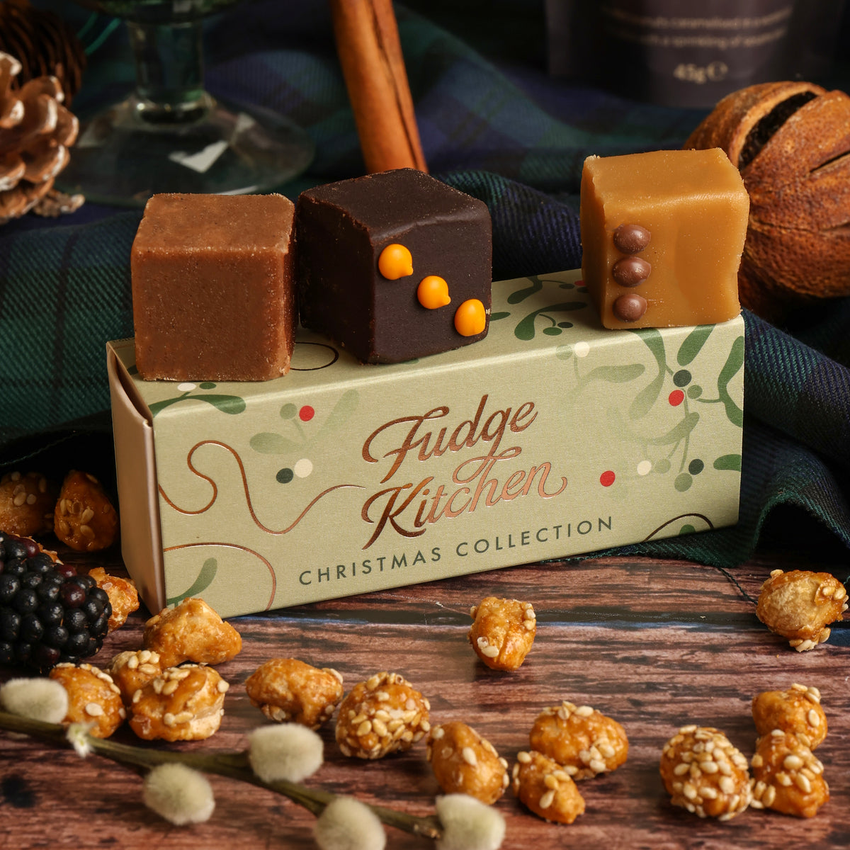 Three decorated fudge cubes displayed on top of a Fudge Kitchen Christmas Collection gift box, surrounded by other snacks, plaid fabric, and festive decorations on a rustic wooden table.