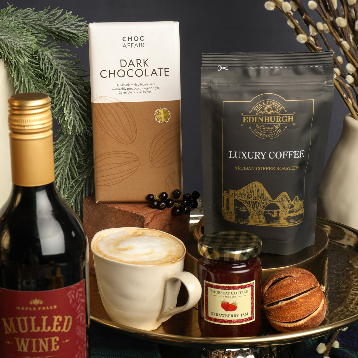 A styled Christmas display on a gold tray featuring a bottle of Maple Falls Mulled Wine, a bar of Choc Affair Dark Chocolate, a bag of Edinburgh Luxury Coffee, a cup of latte coffee, a jar of strawberry jam, and a dried orange, with festive branches and dried flowers in the background.