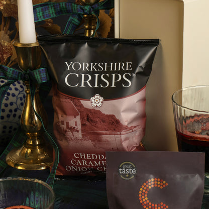 A bag of Yorkshire Crisps Cheddar & Caramelised Onion crisps stands on a tabletop with a candle holder, patterned fabric, and a glass of mulled wine in an indoor setting.