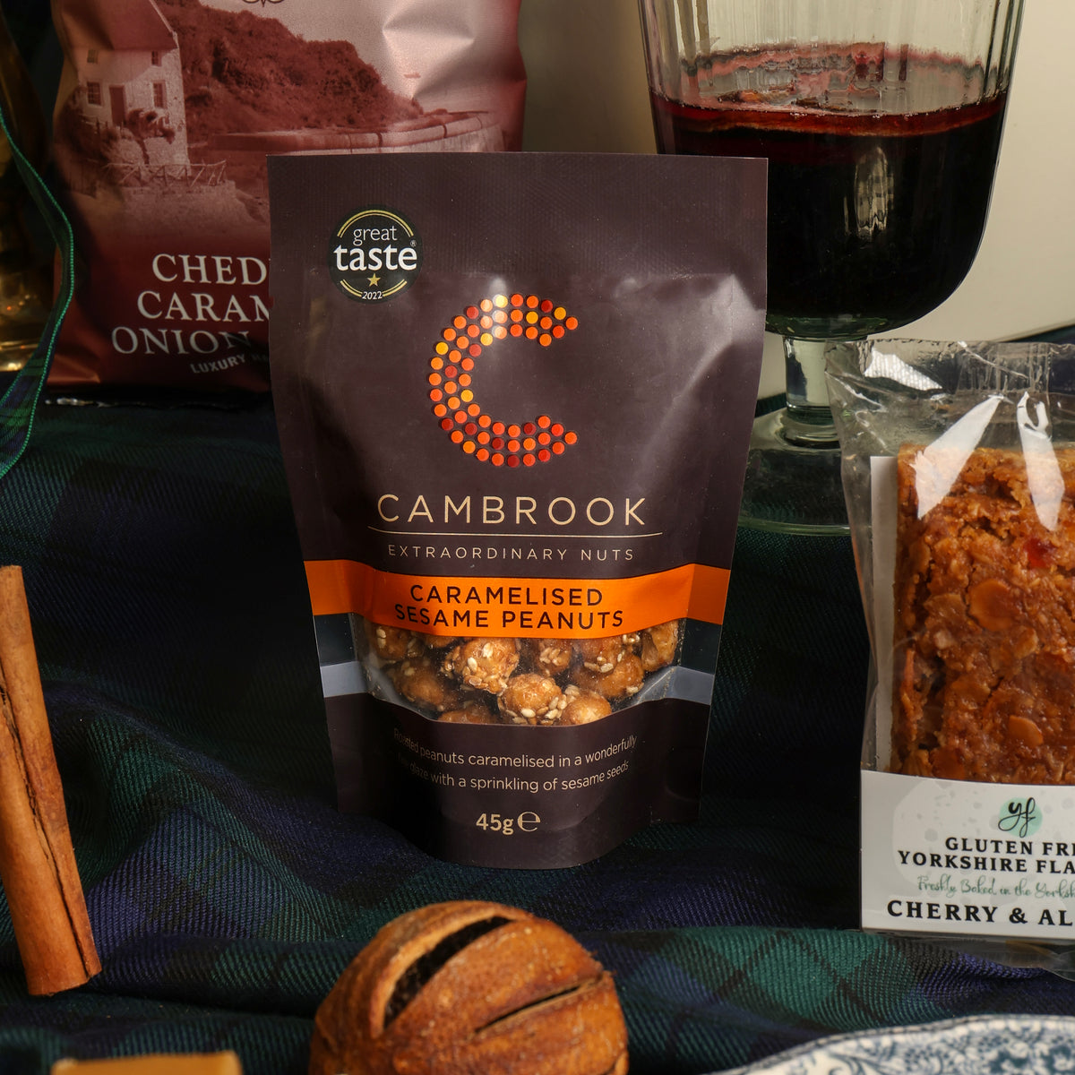 A bag of Cambrook caramelised peanuts is displayed on a dark blue plaid fabric alongside other food items, including a glass of mulled wine, a flapjack, and a dried cinnamon stick decoration.