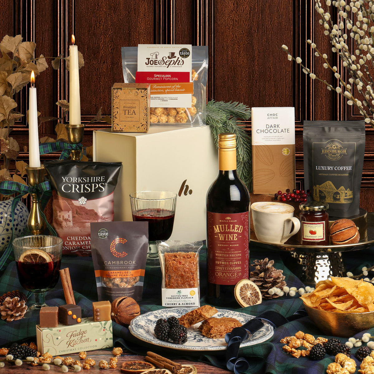 A Christmas gift hamper arrangement featuring an assortment of gourmet snacks from the Luxury Festive Flavours Gift Box, including chocolates, tea, and a bottle of Mulled Wine on a dark tablecloth, styled with candles, seasonal foliage, nuts, and dried flowers.