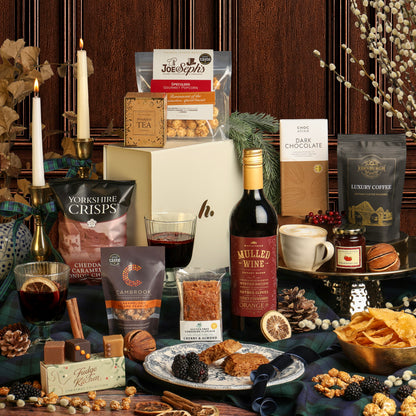 A Christmas gift hamper arrangement featuring an assortment of gourmet snacks from the Luxury Festive Flavours Gift Box, including chocolates, tea, and a bottle of Mulled Wine on a dark tablecloth, styled with candles, seasonal foliage, nuts, and dried flowers.