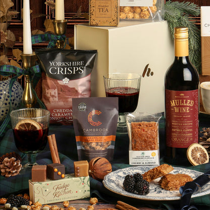 The Luxury Festive Flavours Gift Box, presented on dark teal plaid fabric, with the snacks and drinks surrounding the gift box, including mulled wine, chocolate, popcorn, and fudge pieces on top of a small tray.