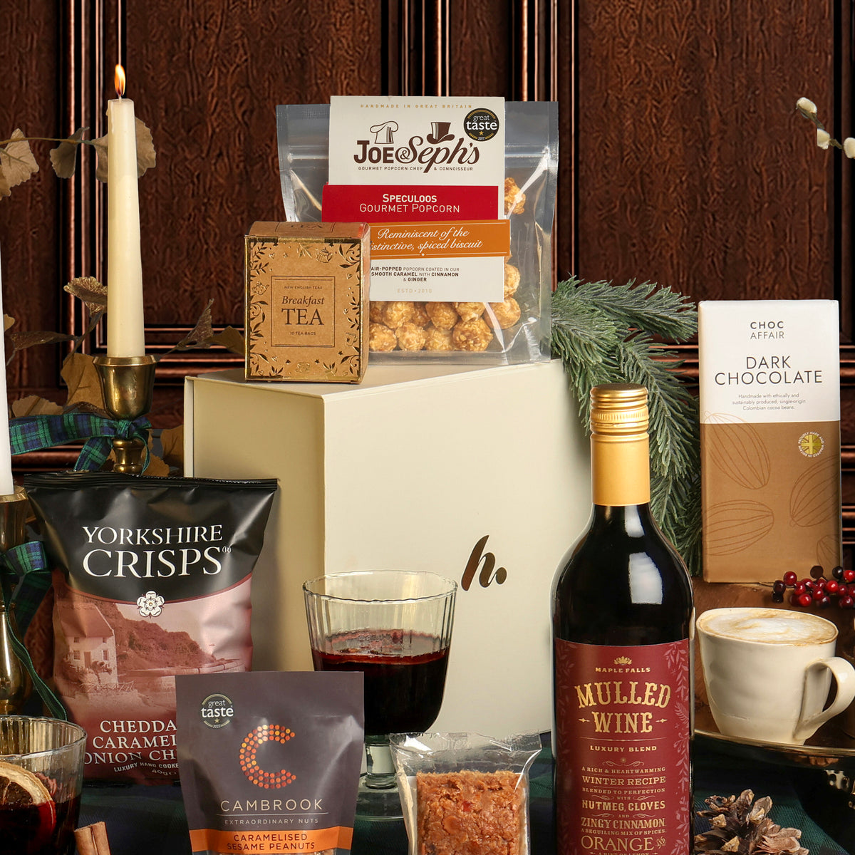 A curated Luxury Festive Flavours Gift Box arrangement with a dark wood panel background, featuring a cream gift box surrounded by snacks, tea, a bottle of mulled wine, styled with a candle and greenery.