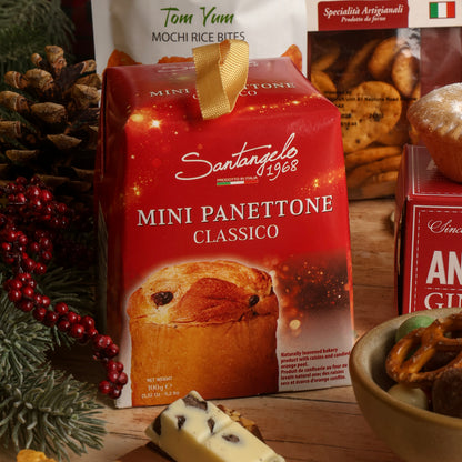 A red box of Santangelo Mini Panettone Classico, with Tom Yum Mochi Rice Bites in the background, styled on a wooden table with pine branches, red berries, pretzels, and chocolate.