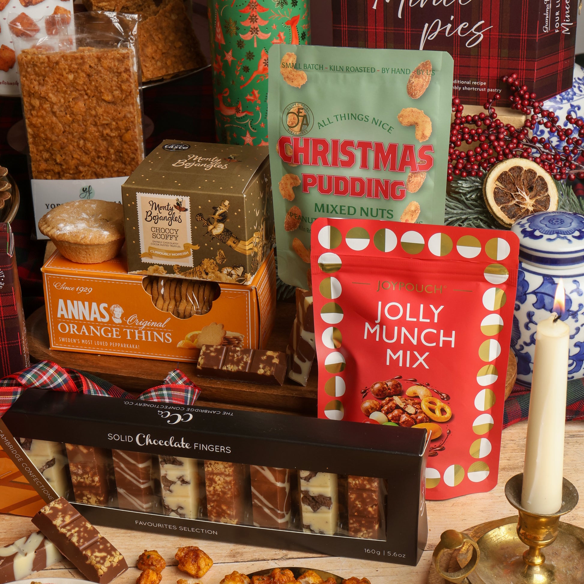 A styled Christmas gift assortment from the Christmas Party Sharing Box, featuring assorted chocolates, mixed nuts, and festive treats. Visible items include a Jolly Munch Mix, Annas Original Orange Thins, Solid Chocolate Fingers, and Christmas Pudding mixed nuts, arranged with plaid textiles, a candle, dried citrus, and berries on a tabletop.