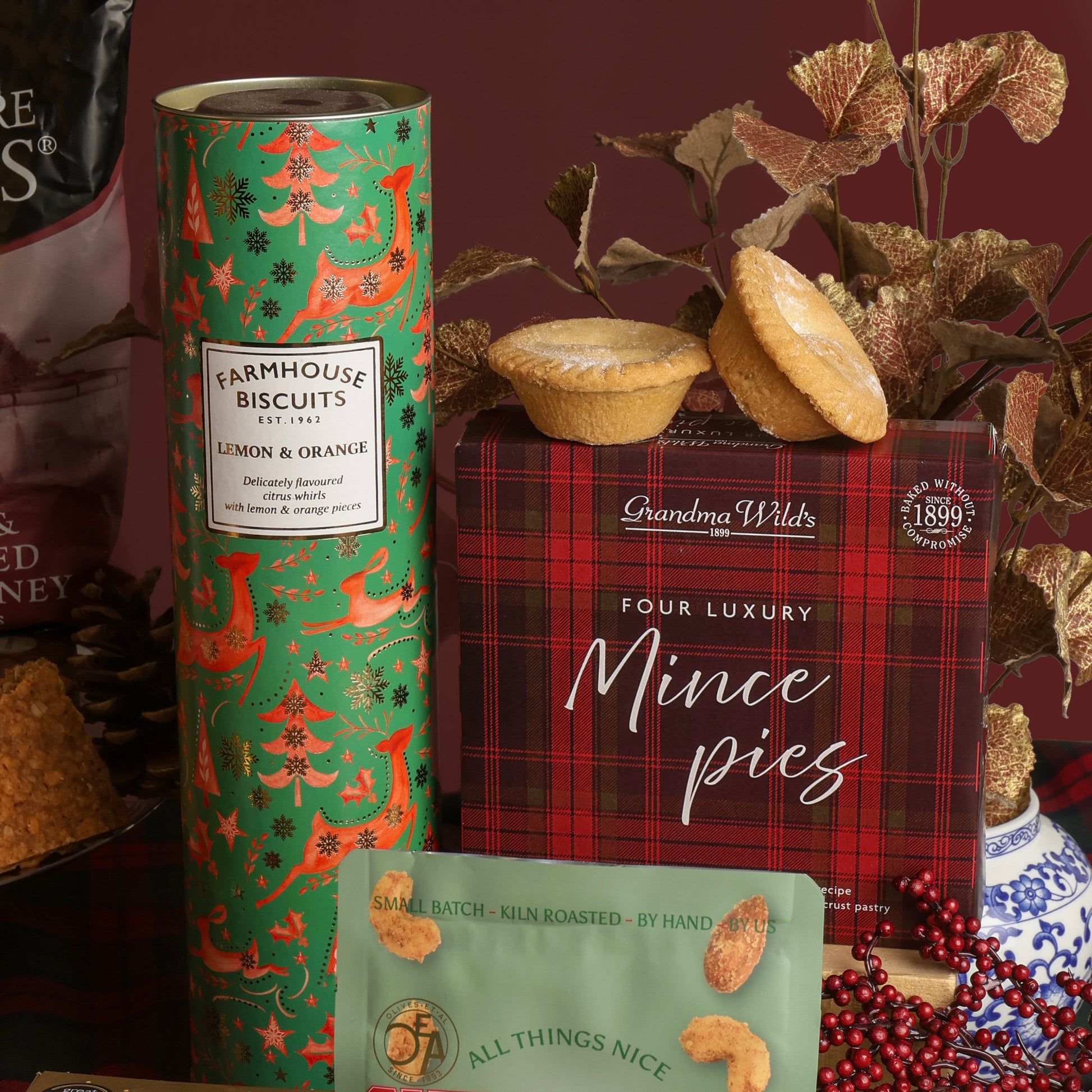 A styled product display featuring a green tube of Farmhouse Biscuits Lemon & Orange, a red plaid box of Grandma Wild's Four Luxury Mince pies, with two mince pies on top of the box, adorned with festive decorations and foliage on a dark red background.