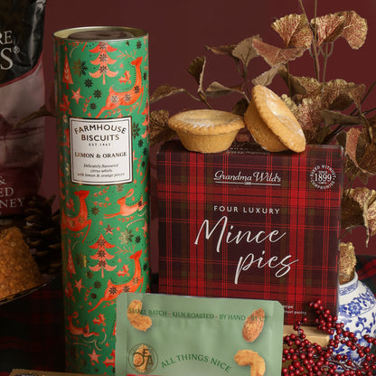 A styled product display featuring a green tube of Farmhouse Biscuits Lemon & Orange, a red plaid box of Grandma Wild's Four Luxury Mince pies, with two mince pies on top of the box, adorned with festive decorations and foliage on a dark red background.