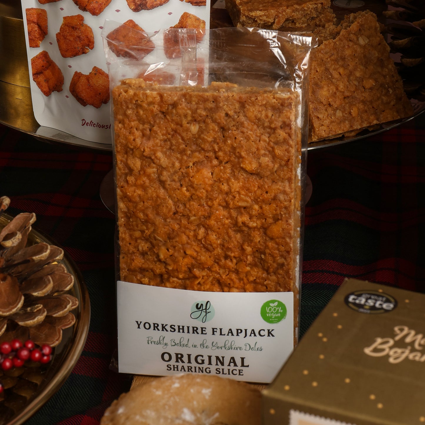 A package of Yorkshire Flapjack original sharing slice, shown in a clear wrapper with the textured golden-brown baked product visible. The background features Christmas props.