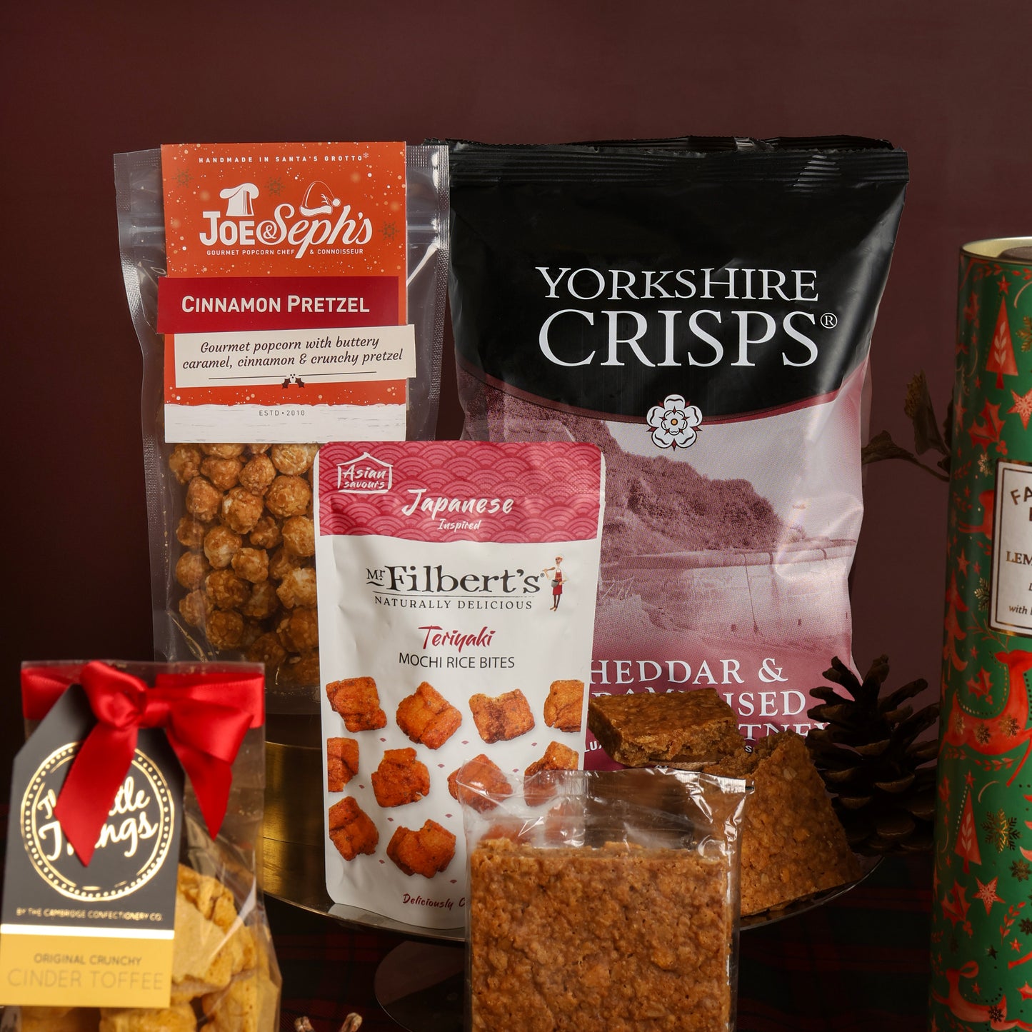 A gift assortment of snacks and treats from the Christmas Party Sharing Box arranged together on a decorative stand against a dark plum background. Visible products include a bag of Joe & Seph's Cinnamon Pretzel Popcorn, a large bag of Yorkshire Crisps, a bag of Mr Filbert's Teriyaki Mochi Rice Bites, and toffee with a red ribbon.