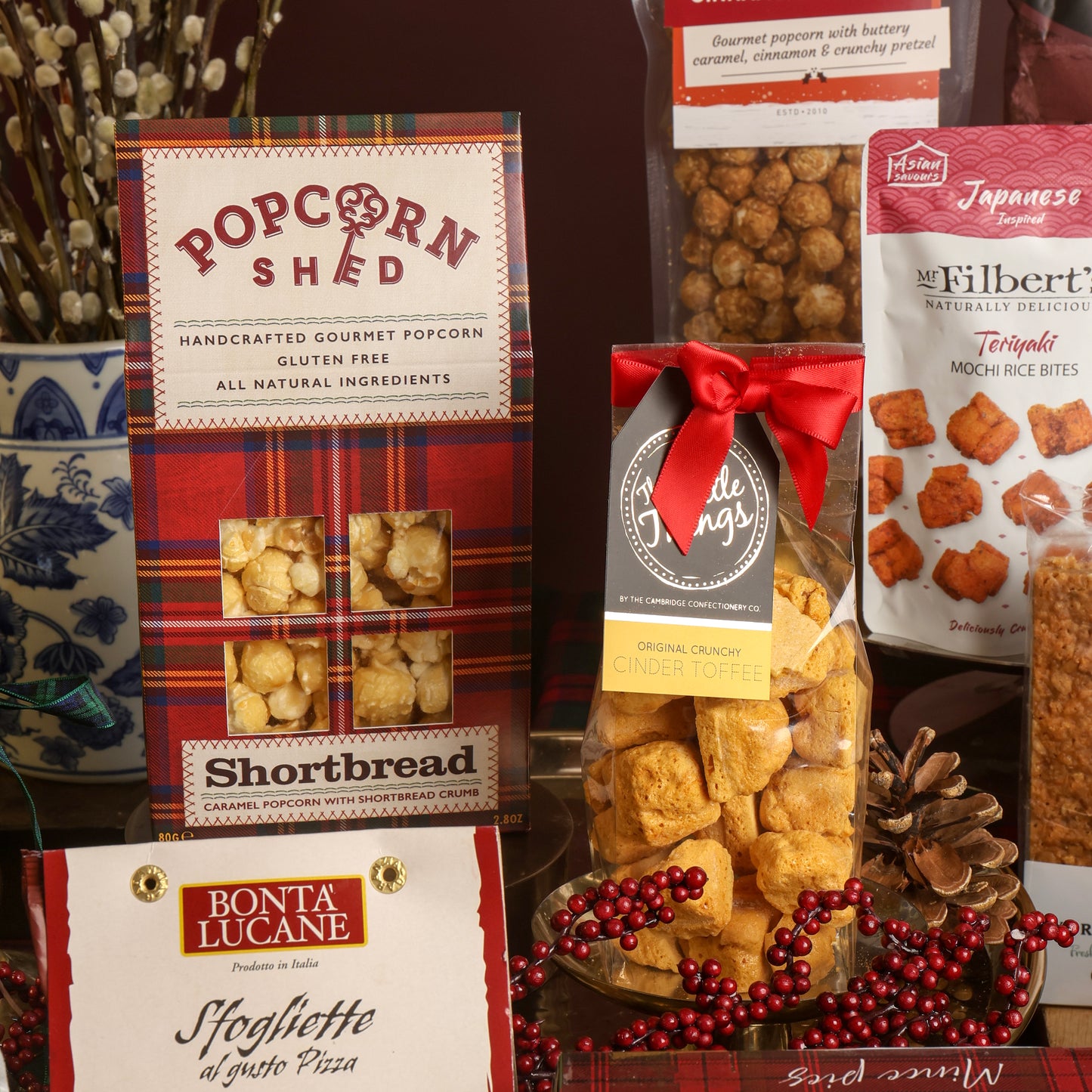 A gift arrangement of gourmet snacks and popcorn, including a red plaid box of Popcorn Shed Shortbread Popcorn, a bag of Mochi Rice Bites, and a clear bag of toffee tied with a red ribbon.