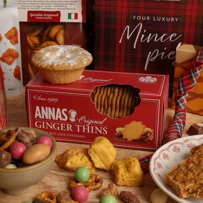 A styled display of Christmas food and treats including a red box of Annas Original Ginger Thins with a mince pie on top of the box, surrounded by a box of mince pies, nuts, and Christmas decorations a wooden surface.