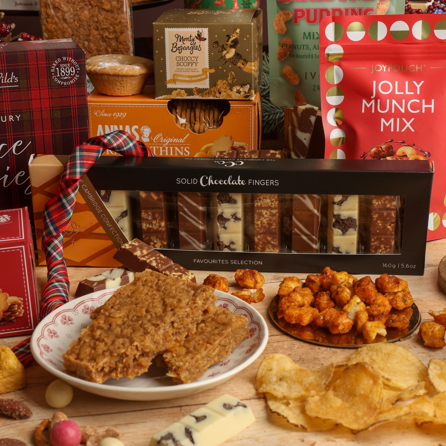 A festive assortment of artisanal treats and snacks arranged on a wooden table, featuring a box of Solid Chocolate Fingers in the centre, surrounded by a Jolly Munch Mix pouch, biscuits, and other Christmas chocolate and flapjacks.