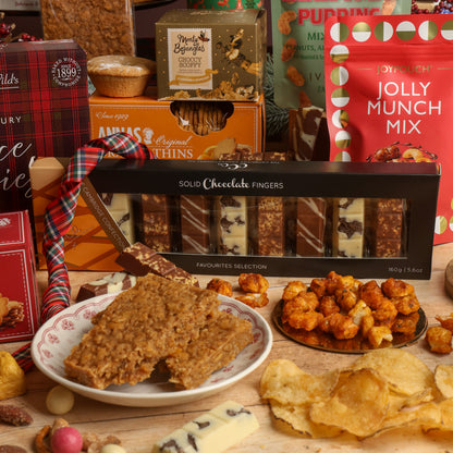 A festive assortment of artisanal treats and snacks arranged on a wooden table, featuring a box of Solid Chocolate Fingers in the centre, surrounded by a Jolly Munch Mix pouch, biscuits, and other Christmas chocolate and flapjacks.