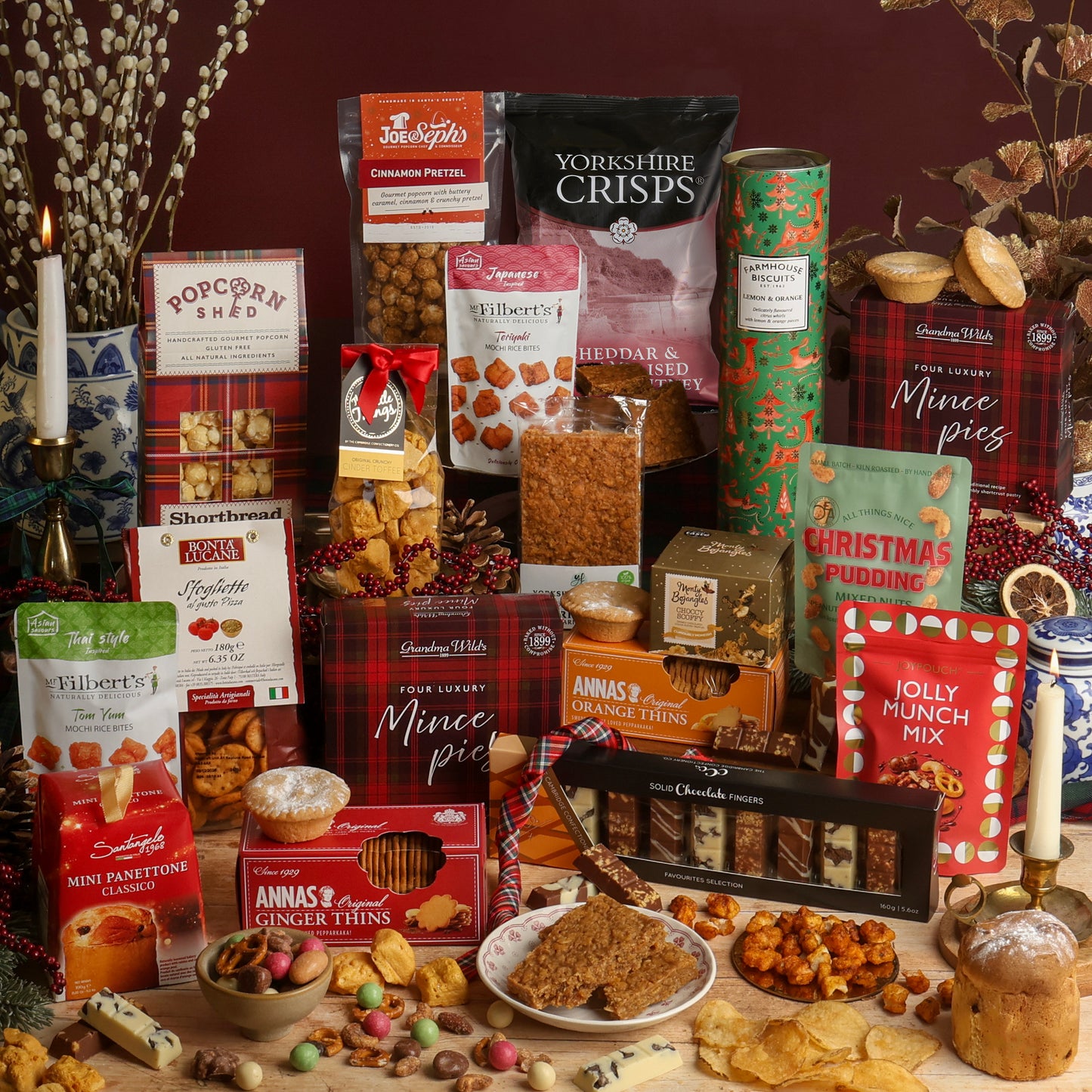 An assortment of food and treats from the Christmas Party Sharing Box hamper, including chocolates, biscuits, and savoury snacks, arranged together on a tabletop with a candle, dried flowers, and Christmas decorations. Visible products include crisps, mince pies, Annas's Orange Thins, Jolly Munch Mix, Christmas Panettone, and various boxed sweets including Popcorn Shed popcorn, toffee, and truffles.