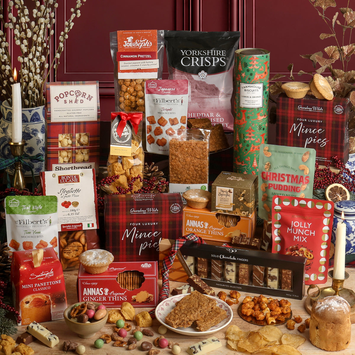 An assortment of food and treats from the Christmas Party Sharing Box hamper, including chocolates, biscuits, and savoury snacks, arranged together on a tabletop with a candle, dried flowers, and Christmas decorations. Visible products include crisps, mince pies, Annas's Orange Thins, Jolly Munch Mix, Christmas Panettone, and various boxed sweets including Popcorn Shed popcorn, toffee, and truffles.