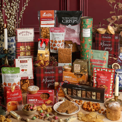 An assortment of food and treats from the Christmas Party Sharing Box hamper, including chocolates, biscuits, and savoury snacks, arranged together on a tabletop with a candle, dried flowers, and Christmas decorations. Visible products include crisps, mince pies, Annas's Orange Thins, Jolly Munch Mix, Christmas Panettone, and various boxed sweets including Popcorn Shed popcorn, toffee, and truffles.
