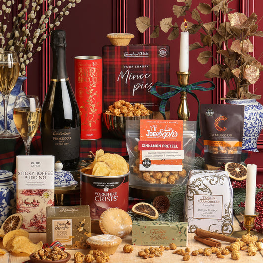 Styled Christmas gifts from the Holly Jolly Christmas Box hamper, including assorted artisanal treats, snacks, and beverages on a rustic wooden surface against a dark red background. Visible items include a bottle of Prosecco, a box of Four Luxury Mince Pies, a bag of Joe & Seph's Cinnamon Pretzel popcorn, a bar of Sticky Toffee Pudding chocolate, a tin of Yorkshire Crisps, a bag of Cambrook nuts, along with candles, glasses of Prosecco, and decorative dried botanicals.