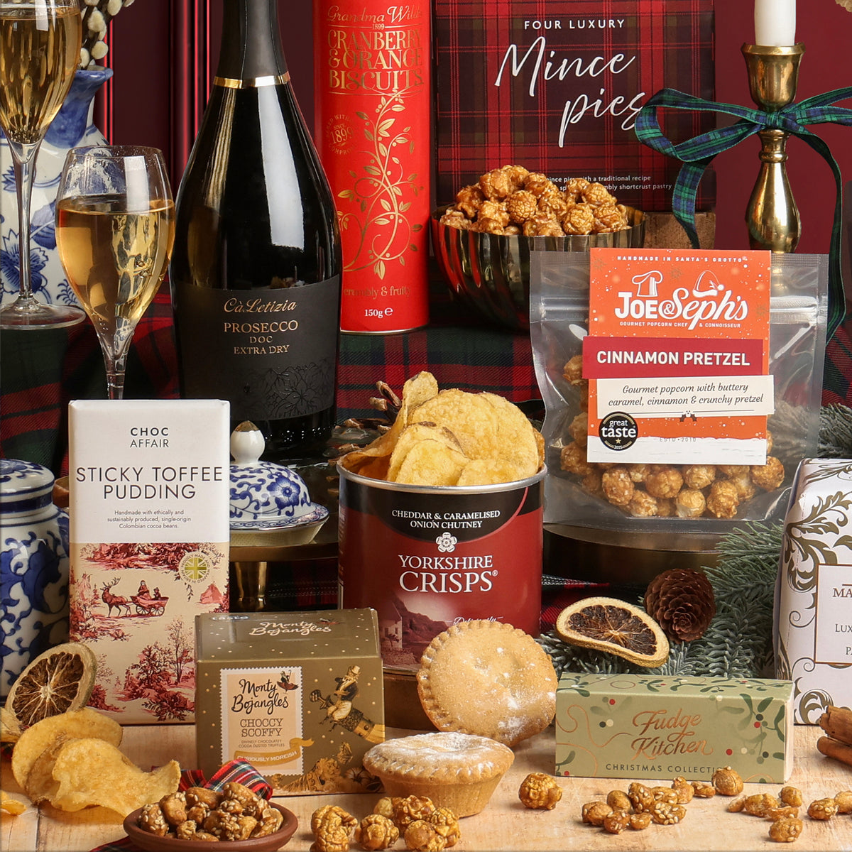 Holly Jolly Christmas Box styled in a festive setting with decorations, plaid ribbon, dried fruit, and Prosecco glasses, showcasing the sweet and savoury snacks included in the hamper. 