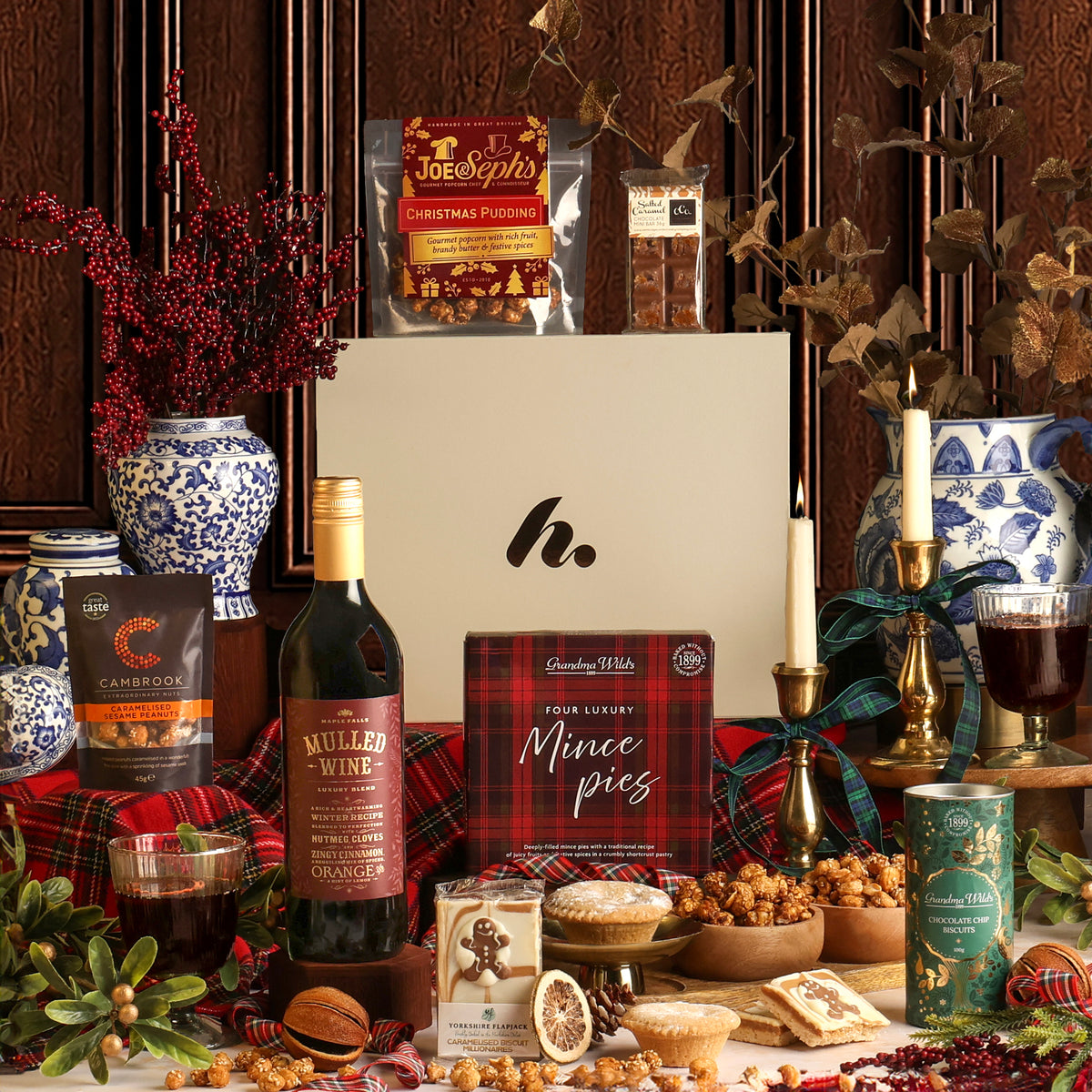 The Festive Night In hamper arrangement featuring a white gift box with a logo, surrounded by Christmas treats and drinks including a bottle of mulled wine, a tin of chocolate chip biscuits, mince pies, nuts, candles, and blue-and-white ceramic vases against a dark wood panel background.