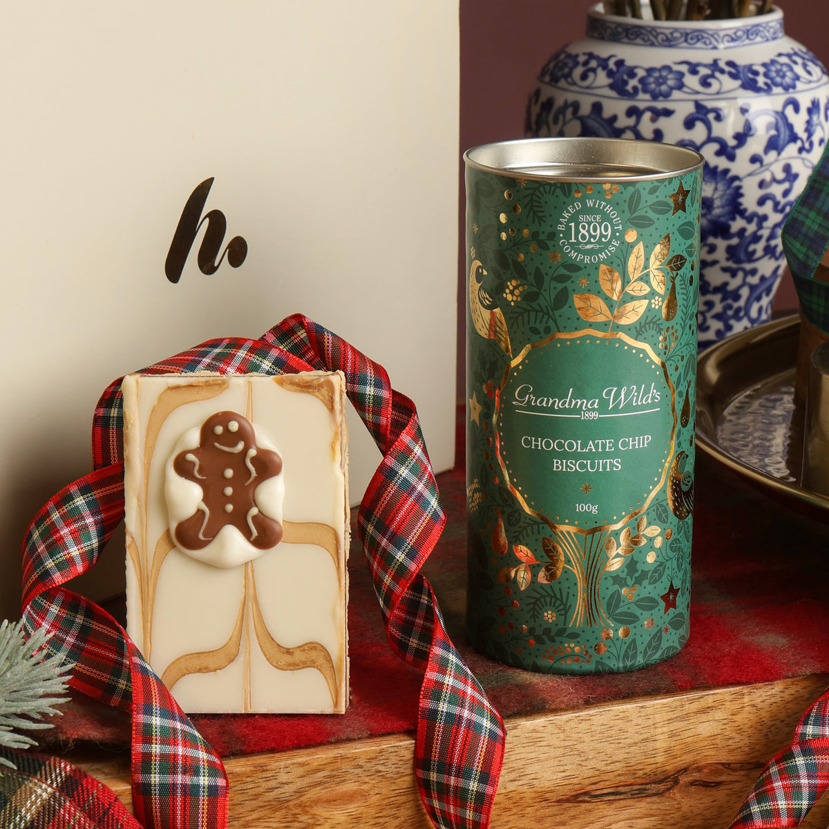 A tin of Grandma Wild's Chocolate Chip Biscuits next to a chocolate slice decorated with a gingerbread person, wrapped in a red plaid ribbon, set on a wooden surface with a blue and white ceramic vase in the background.