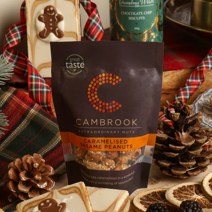 A bag of Cambrook Caramelised Sesame Peanuts placed in a styled Christmas scene with plaid fabric, pinecones, dried orange slices, blackberries, and tray-bake slices. A tin of Grandma Wild's Chocolate Chip Biscuits is visible in the background.