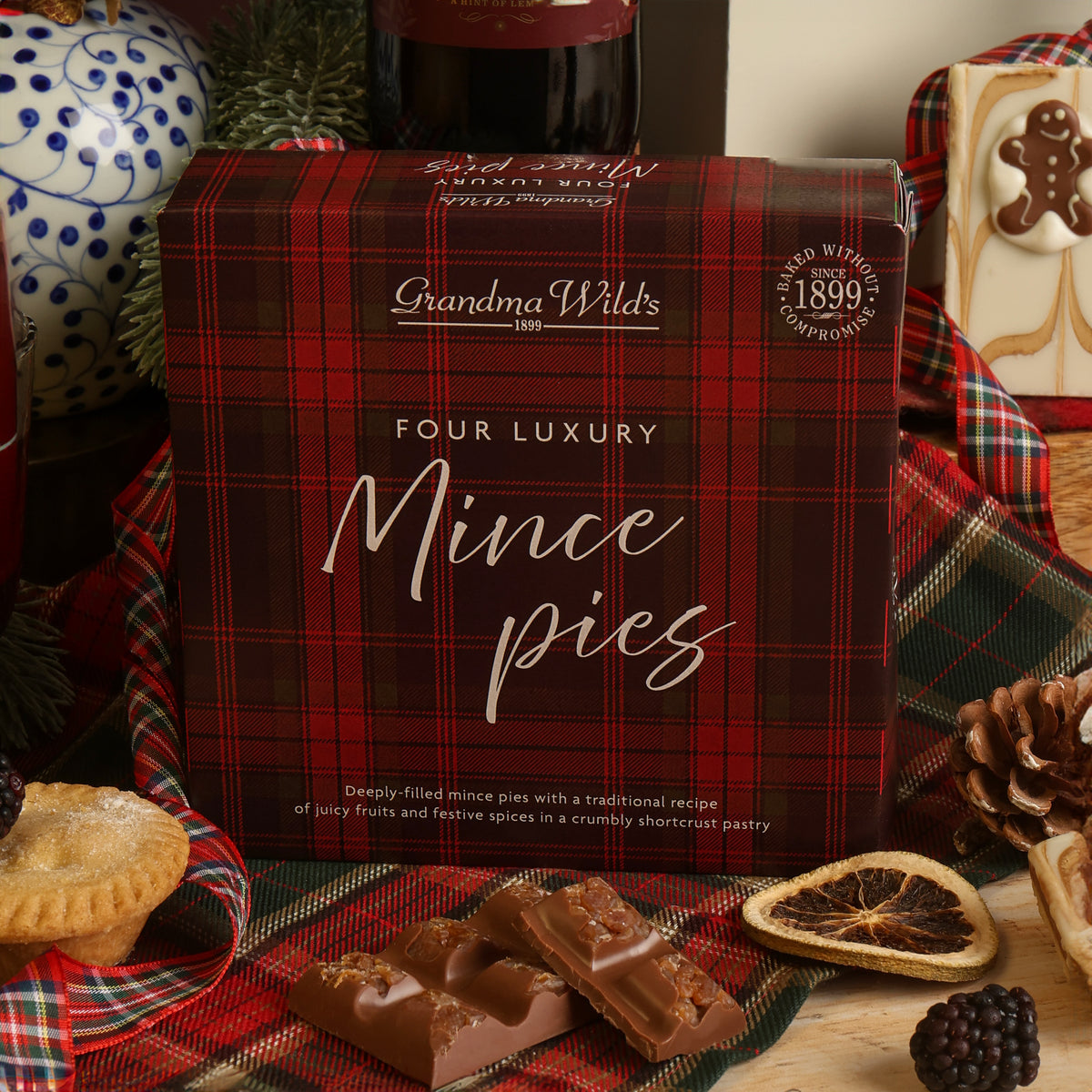A box of Grandma Wild's Four Luxury Mince Pies, styled on a festive Christmas table with chocolate, a tray-bake slice decorated with a gingerbread person, dried orange slices, and plaid fabric.