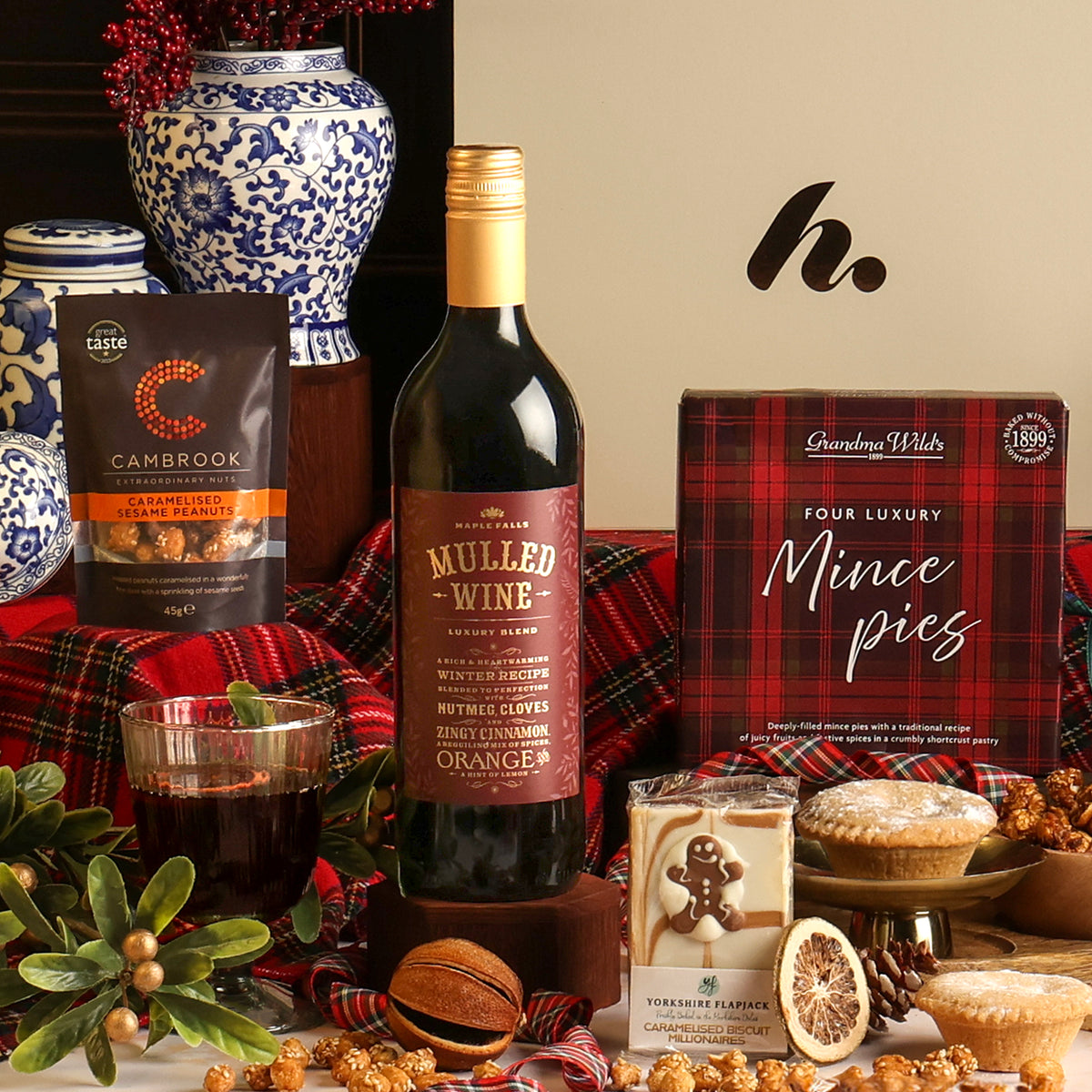 A styled Christmas gift hamper featuring a bottle of Mulled Wine, a bag of Cambrook peanuts, a box of mince pies, and a pack of festive flapjack decorated with a gingerbread person, arranged on a red plaid cloth with decorative ceramic vases, greenery, and popcorn.