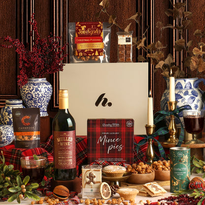 The Festive Night In hamper arrangement featuring a white gift box with a logo, surrounded by Christmas treats and drinks including a bottle of mulled wine.