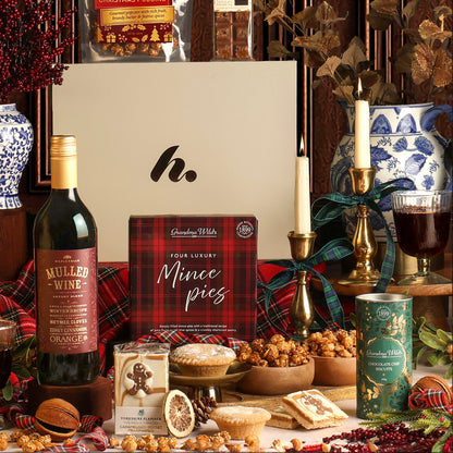 The Festive Night In Christmas hamper with its contents displayed, including a bottle of mulled wine, a box of mince pies, a tin of biscuits and popcorn, styled with dried fruit, candles and plaid fabric in a Christmas setting.