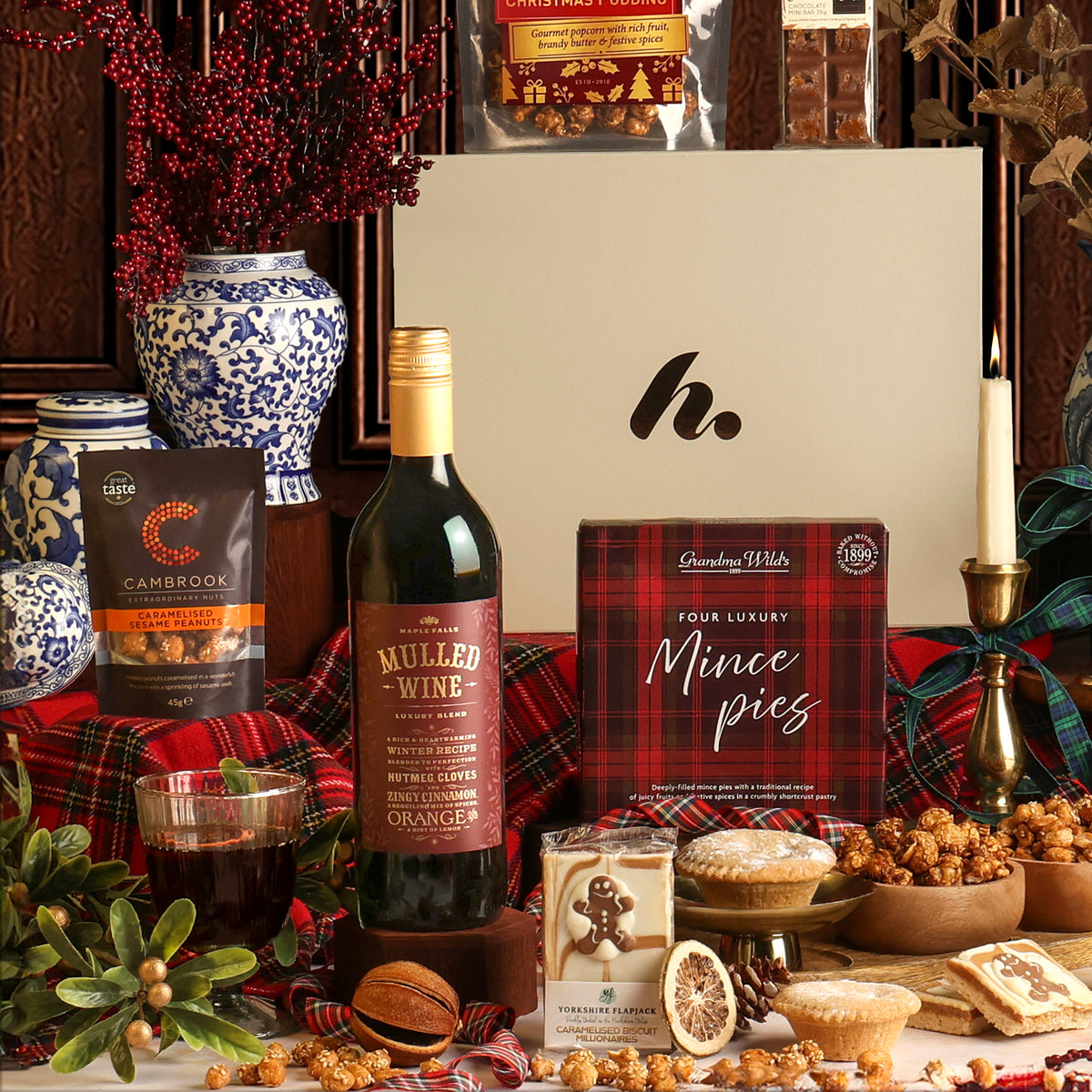 A festive Christmas gift hamper featuring a bottle of Mulled Wine, a box of Luxury Mince Pies, and other Christmas treats arranged on a red plaid surface in front of a large cream gift box.