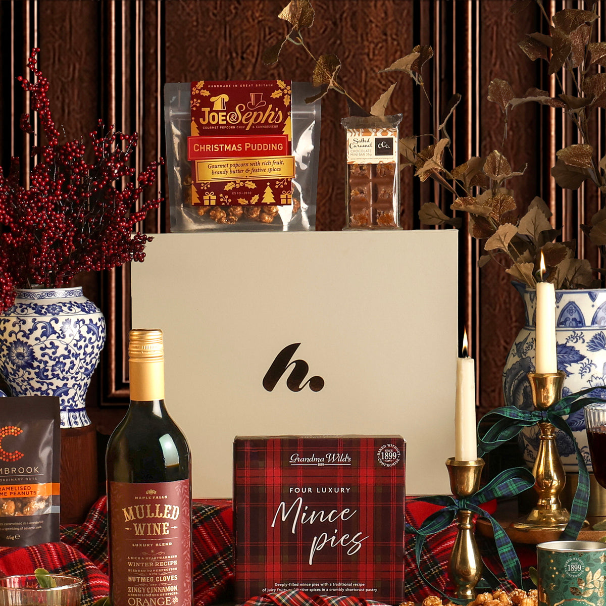 The Festive Night In Christmas hamper arrangement featuring a white gift box surrounded by gourmet treats including mince pies, a bottle of mulled wine, and other seasonal items on a red plaid fabric surface.