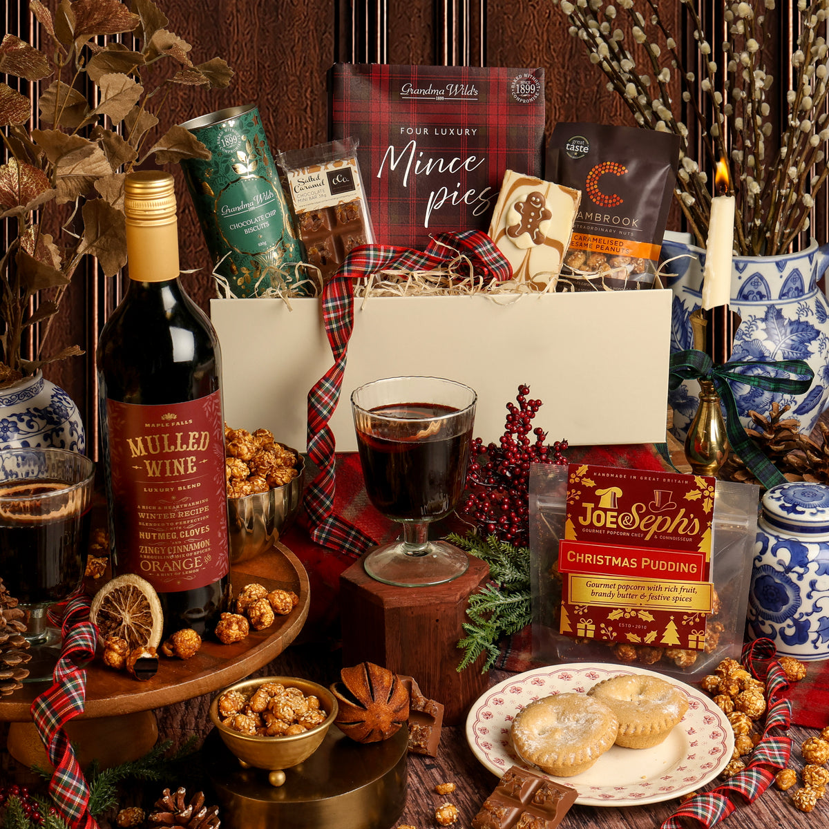 The Festive Night In gift hamper arranged on a rustic wooden surface, featuring a bottle of mulled wine, a box of mince pies, Christmas Pudding popcorn, and other Christmas treats, with a white gift box, plaid ribbon, a candle, and seasonal decorations.