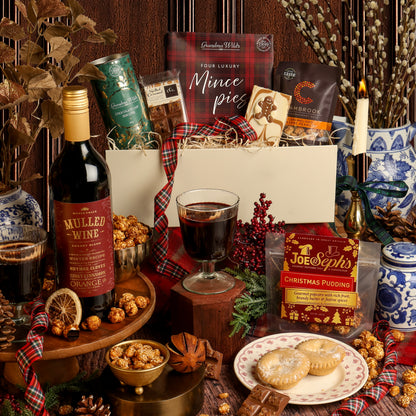 The Festive Night In gift hamper arranged on a rustic wooden surface, featuring a bottle of mulled wine, a box of mince pies, Christmas Pudding popcorn, and other Christmas treats, with a white gift box, plaid ribbon, a candle, and seasonal decorations.