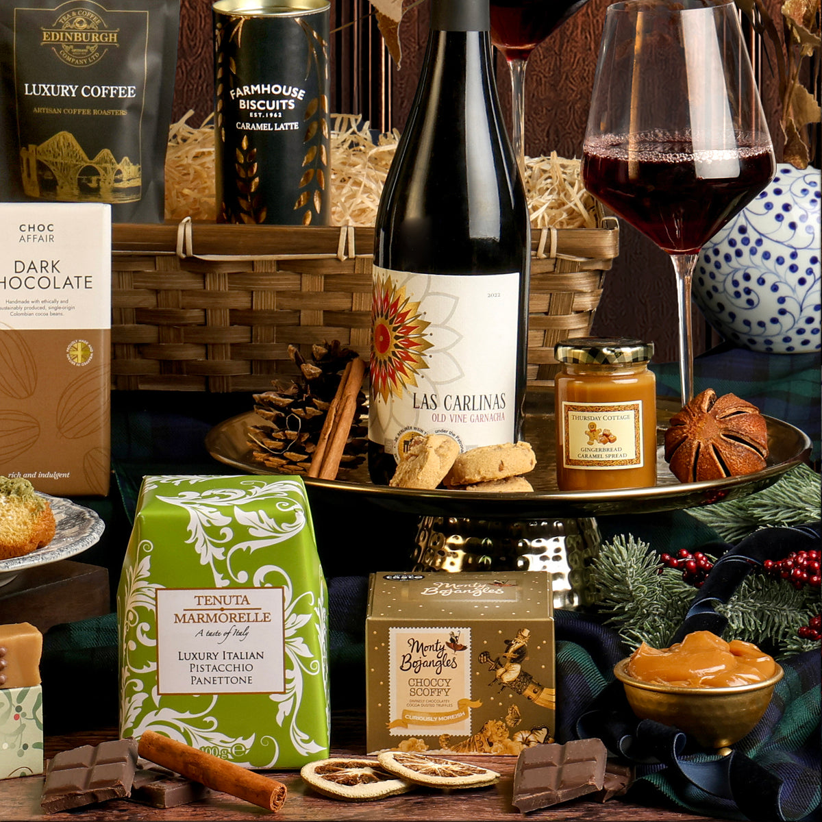 The Gold Standard Christmas Hamper featuring a bottle of red wine, a green box of panettone, a box of chocolate truffles, chocolate, biscuits, and glasses of red wine styled on a dark wood panelled background.