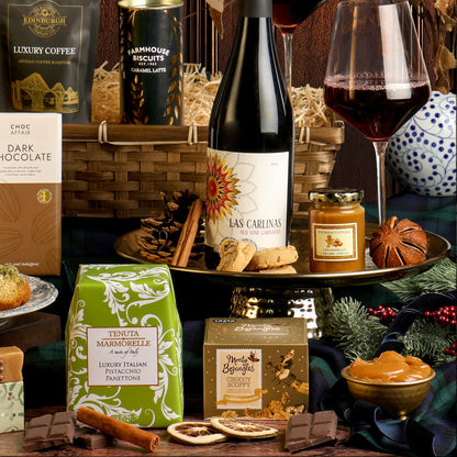The Gold Standard Christmas Hamper featuring a bottle of red wine, a green box of panettone, a box of chocolate truffles, chocolate, biscuits, and glasses of red wine styled on a dark wood panelled background.