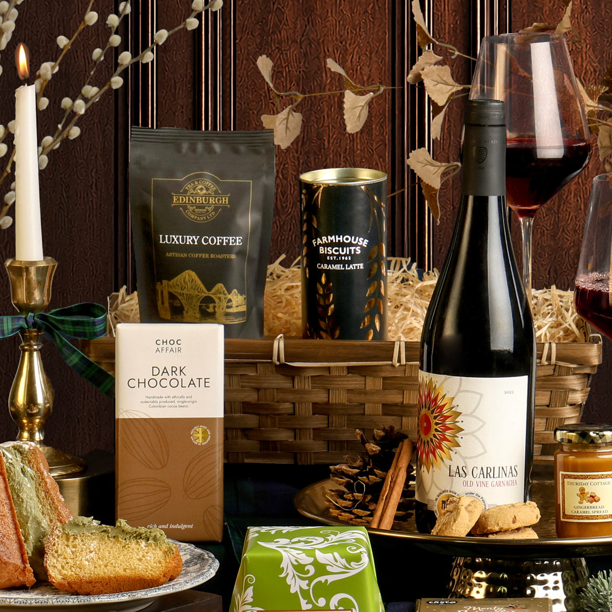 The Gold Standard Christmas Hamper arranged to display the contents, including a bottle of red wine, a bag of Edinburgh Luxury Coffee, a bar of dark chocolate, and biscuits, with a reusable bamboo tray, set against a dark wood panel background.