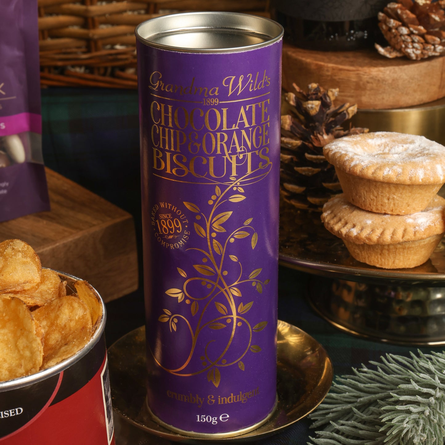 A purple tin of Grandma Wild's Chocolate Chip & Orange Biscuits stands on a gold tray, surrounded by Christmas props including a plate of stacked mince pies, pinecones, and a bowl of crisps.