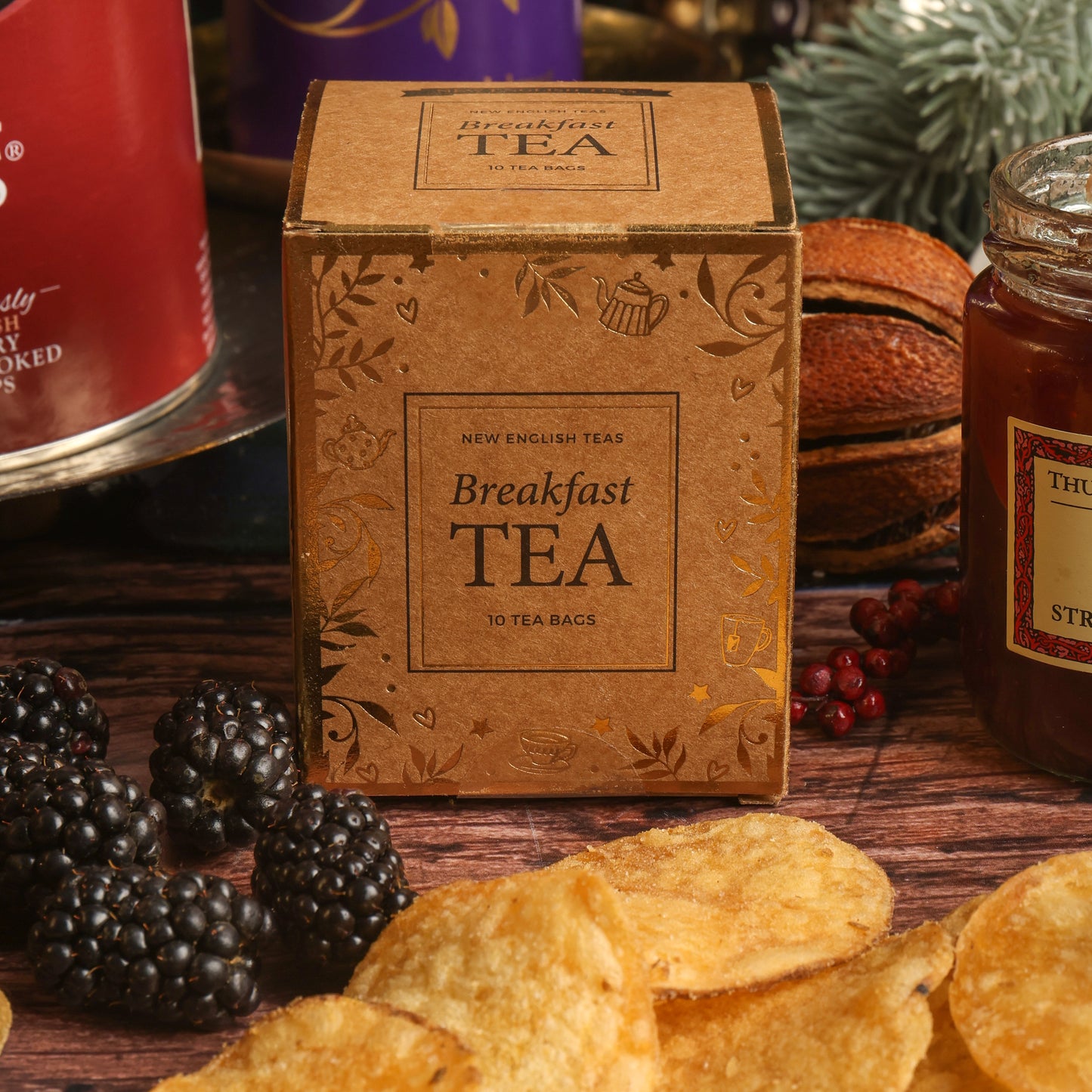 A paper box of Breakfast Tea centred on a rustic wooden table, surrounded by Christmas themed props including blackberries and dried citrus, along with scattered crisps, and a jar of jam.