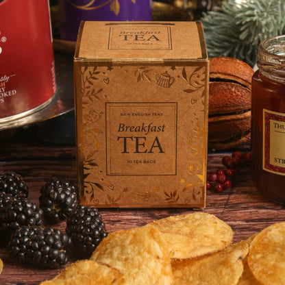 A paper box of Breakfast Tea centred on a rustic wooden table, surrounded by Christmas themed props including blackberries and dried citrus, along with scattered crisps, and a jar of jam.