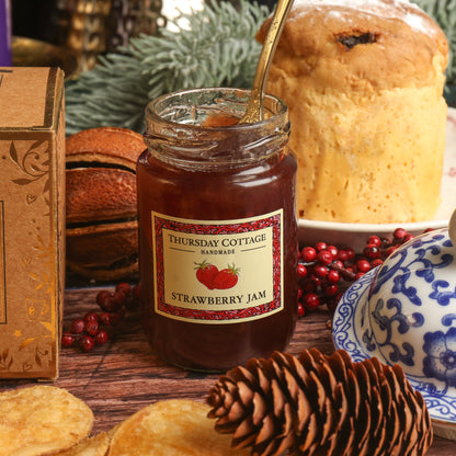 A glass jar of Thursday Cottage Strawberry Jam sits on a wooden table, in front of a panettone, surrounded by Christmas props including red berries, a pinecone, and a decorative box of tea.