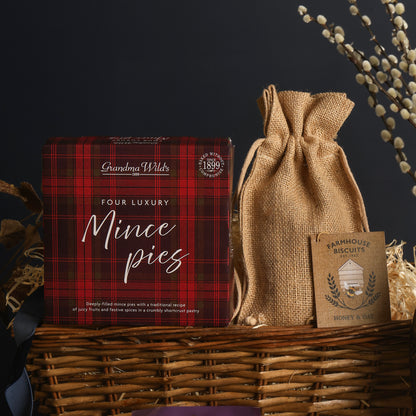 A gift basket arrangement from the Winter Wonders Christmas Gift Basket, featuring a red tartan box of luxury mince pies, and a natural hessian drawstring pouch from Farmhouse Biscuits, in a wicker basket against a dark background.