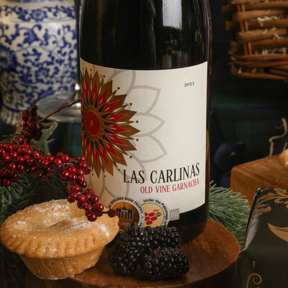 A bottle of Las Carlinas Garnacha red wine is displayed on a wooden surface with a mince pie, blackberries, and festive greenery in a styled setting.