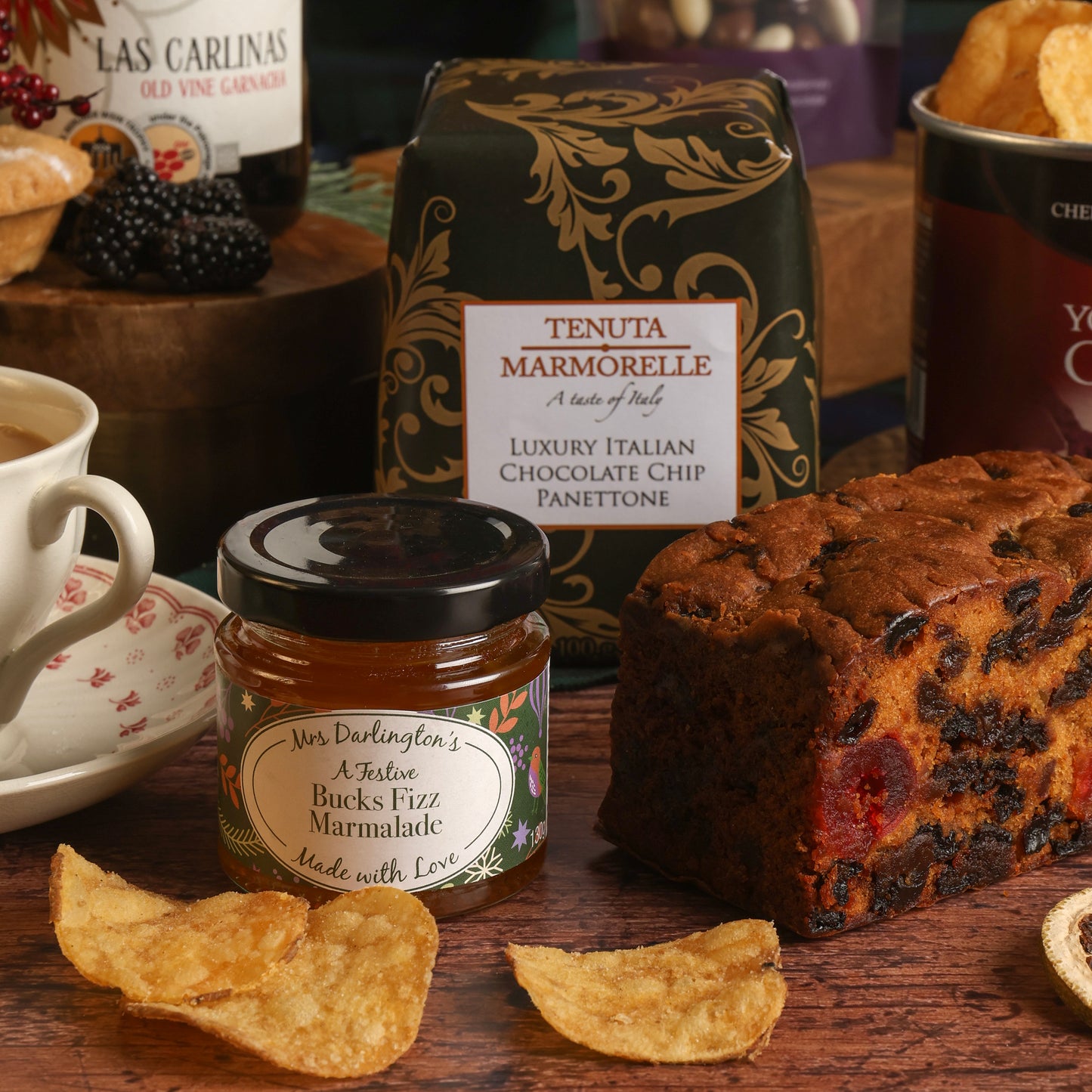 A styled tea and snack scene on a wooden table with a bag of Tenuta Marmorelle Italian chocolate chip panettone, a jar of Bucks Fizz Marmalade, a loaf of fruit cake, and crisps scattered in front.
