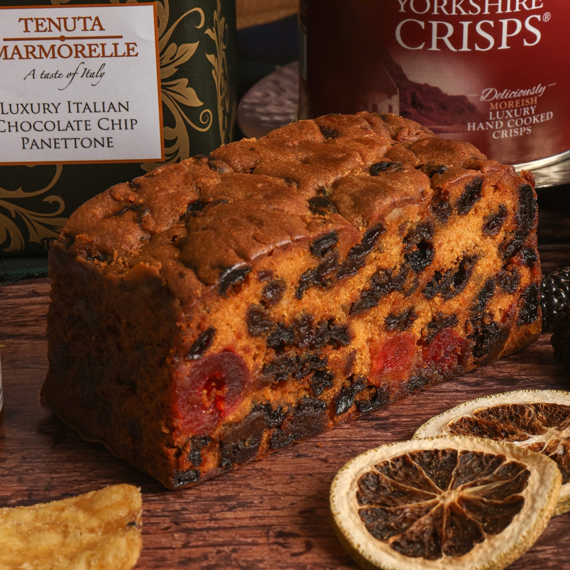 A loaf of fruit cake, rich with fruit pieces, displayed on a rustic wooden surface, with a Tenuta Marmorelle luxury chocolate chip panettone box in the background, along with crisps and dried citrus decorations.
