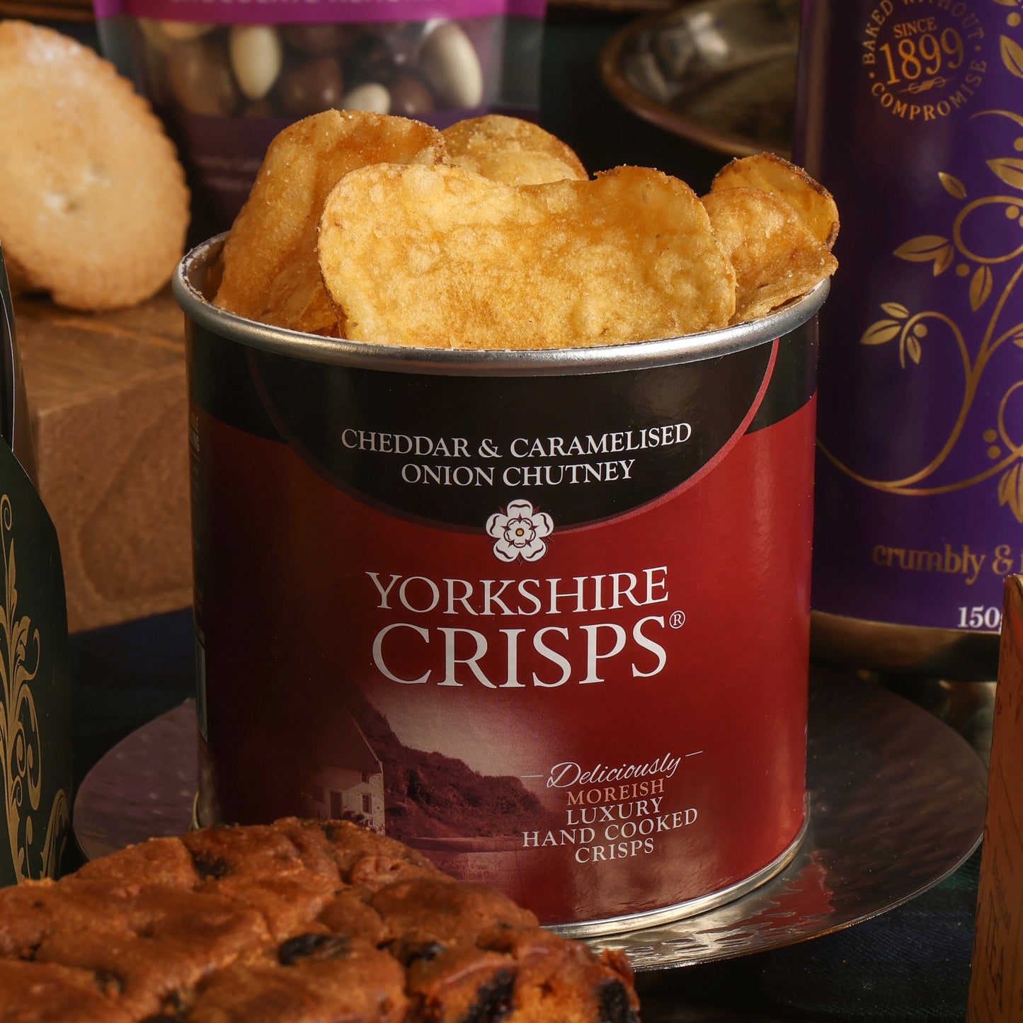 An open tin of Yorkshire Crisps Cheddar & Caramelised Onion Chutney, filled with golden crisps, displayed on a reflective stand with other food items in the background.