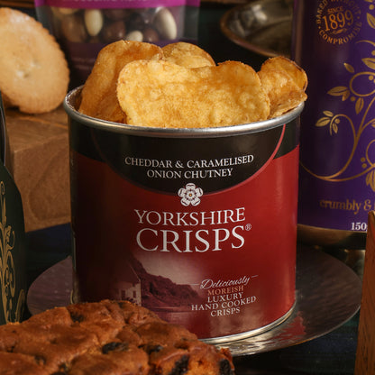 An open tin of Yorkshire Crisps Cheddar & Caramelised Onion Chutney, filled with golden crisps, displayed on a reflective stand with other food items in the background.