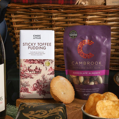 A gift basket arrangement featuring a bag of Cambrook Chocolate Almonds and a bar of Choc Affair Sticky Toffee Pudding chocolate, with a mince pie and crisps in the foreground.